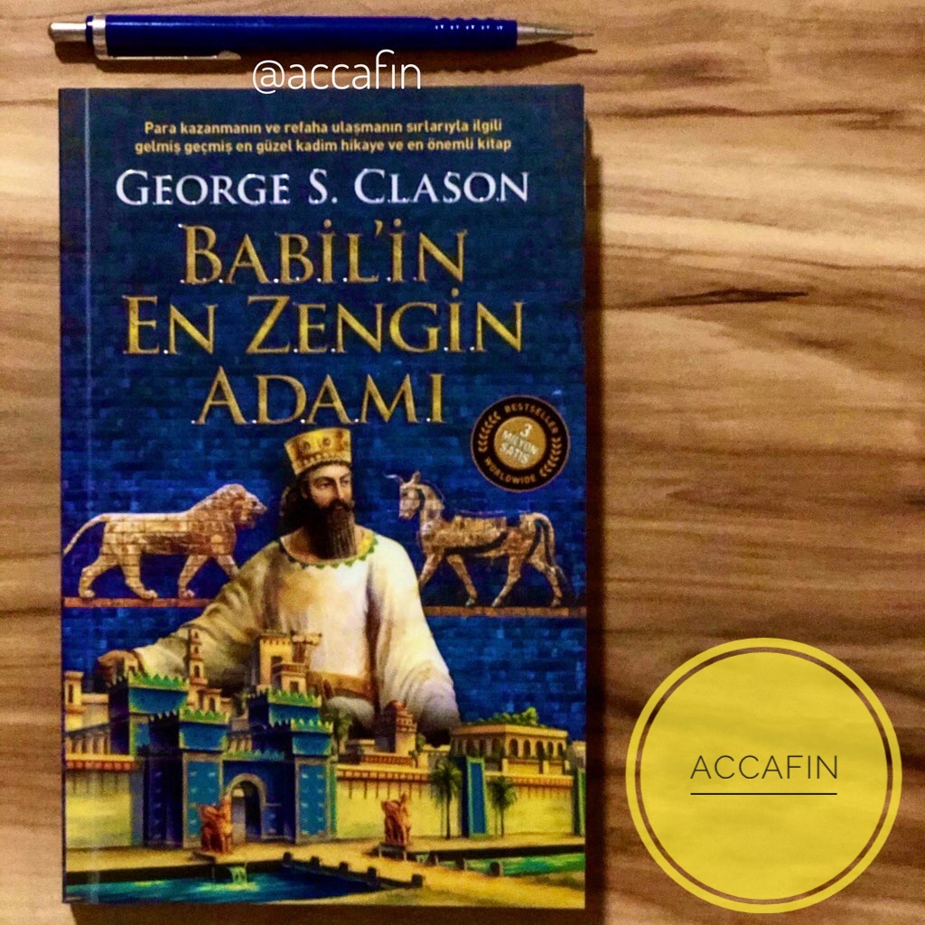 George S. Clason'ın Babil'in En Zengin Adamı kitabı ahşap masada, yanında kalem ve accafin rozeti