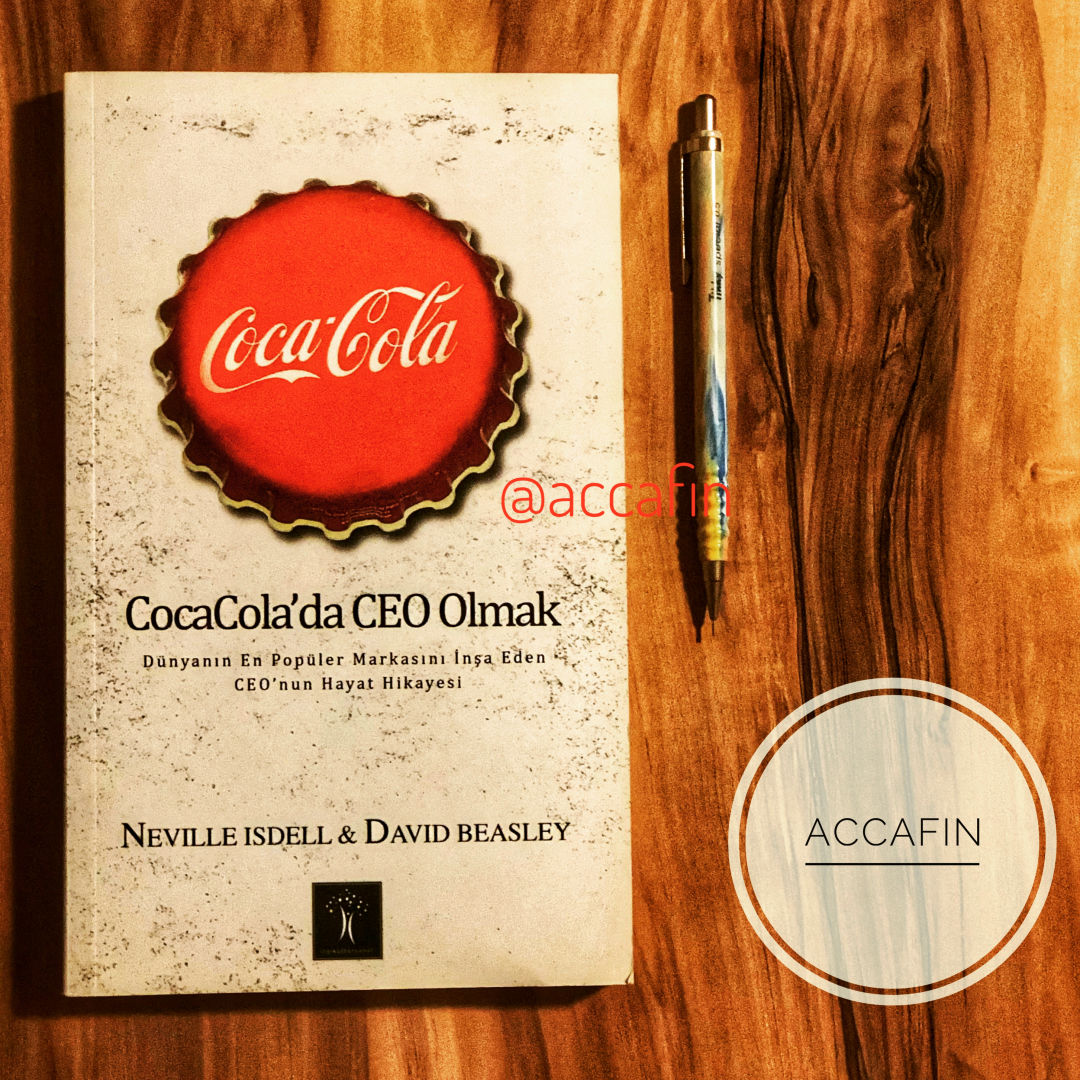 Neville Isdell ve David Beasley'nin Coca-Cola'da CEO Olmak kitabı ahşap masada, yanında kalem ve accafin rozeti